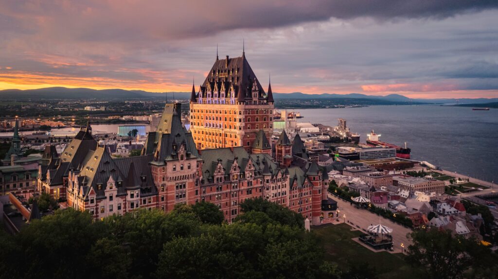 Aerial sunset view of Fairmont Le Château Frontenac's turreted castle-like silhouette overlooking Quebec City and the St. Lawrence River under a dramatic pink-and-gray sky.