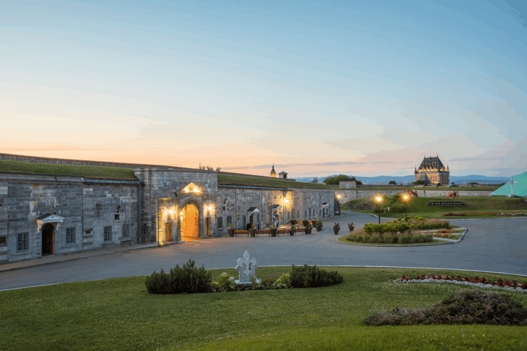 Twilight view of the Citadelle de Québec's illuminated stone fortress entrance, with the Château Frontenac visible in the distance across manicured lawns.
