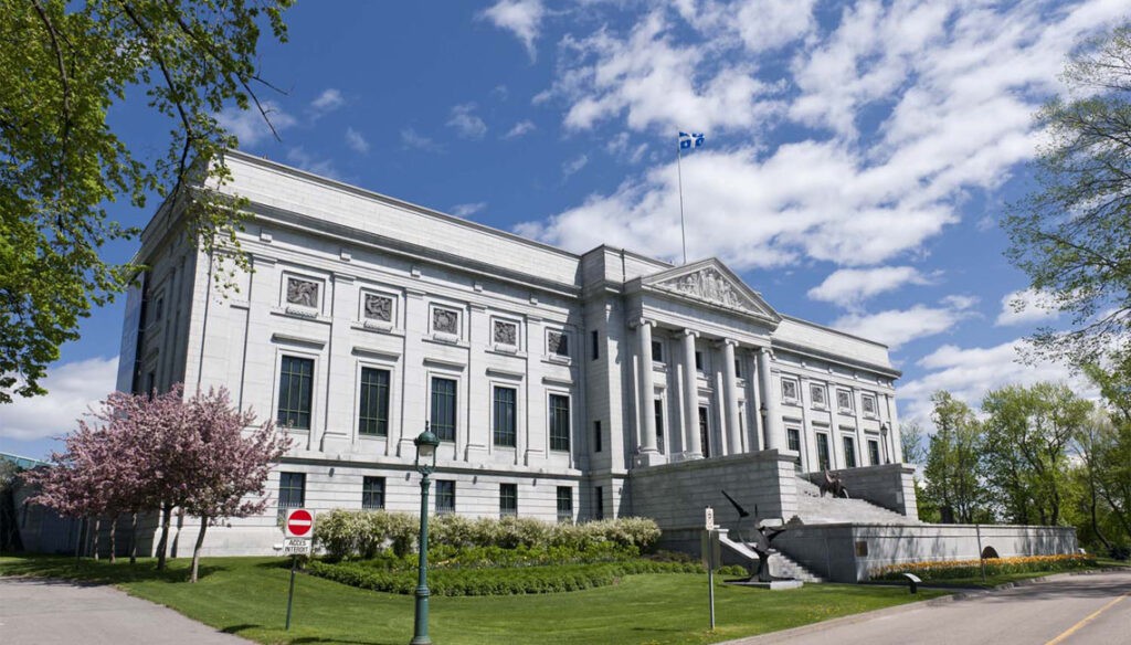 Exterior view of the Musée national des beaux-arts du Québec, a neoclassical stone building with grand columns and a pediment facade, set against a blue sky with blooming trees.