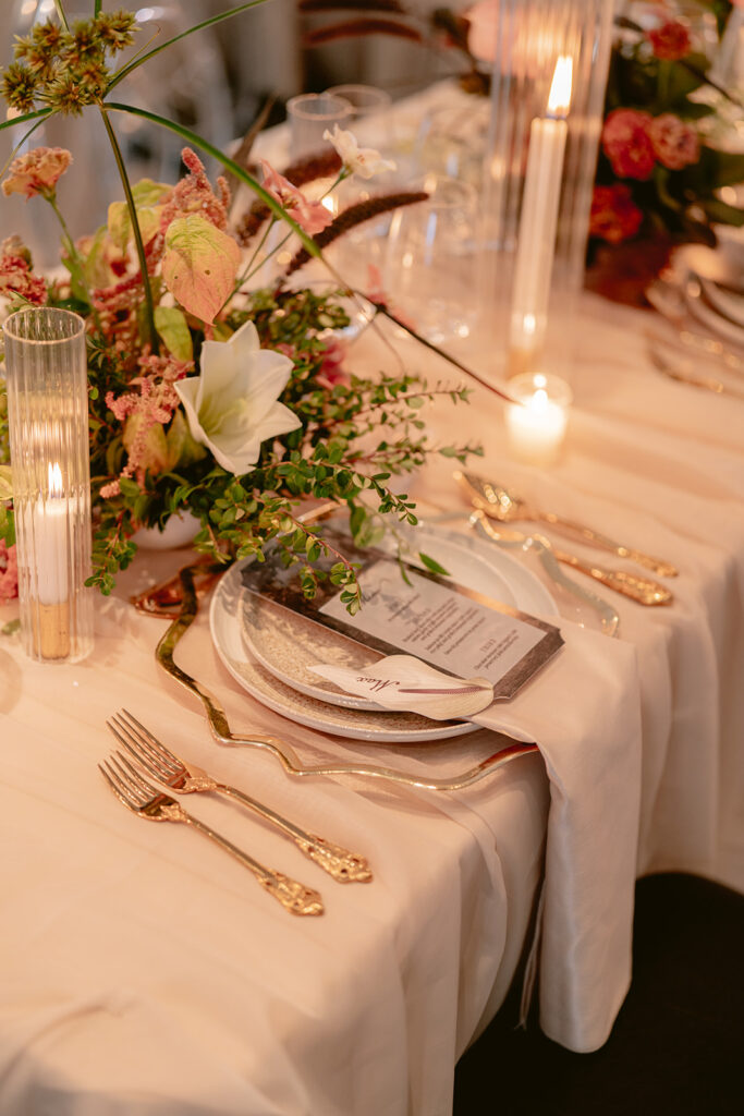 Wedding table setting with gold flatware, glass candle holders, and a wild floral centerpiece in soft pink and burgundy tones, styled by Amelia at A. Carleton Weddings.