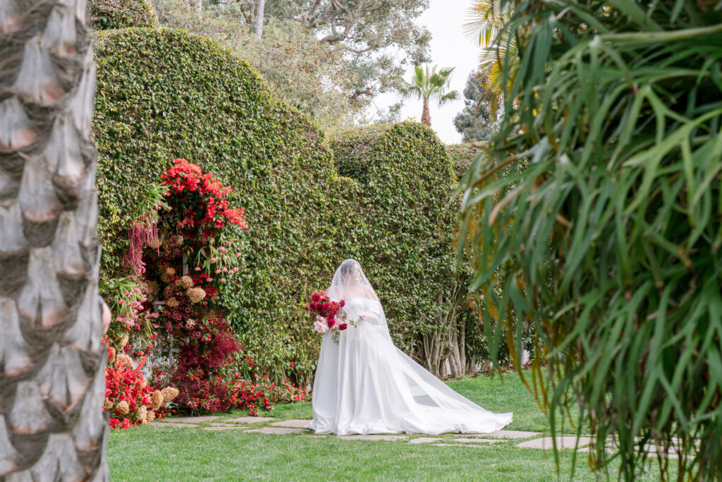 Bride in a veiled gown carrying a bold red bouquet, standing before a vibrant floral archway set against tall manicured hedges and palm trees in a lush garden setting.