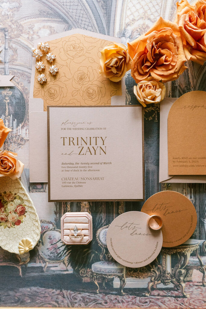 Wedding invitation flat lay in kraft and cream tones, styled with orange roses, a ring box, and vintage-patterned backdrop.