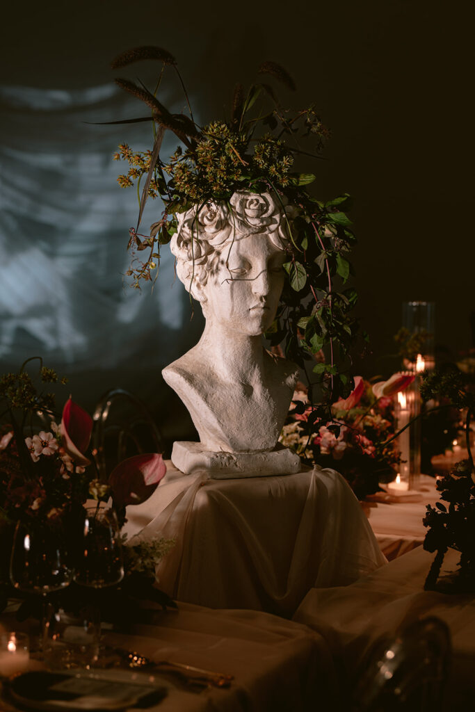 Moody candlelit table featuring a classical bust adorned with a floral crown, surrounded by dark, romantic blooms, styled by Amelia at A. Carleton Weddings.