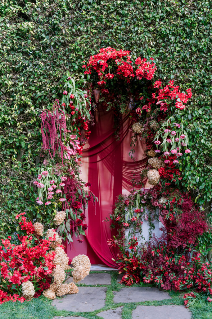 Vibrant floral archway of red and burgundy blooms framing a draped fabric entrance, set into a wall of dense green ivy.