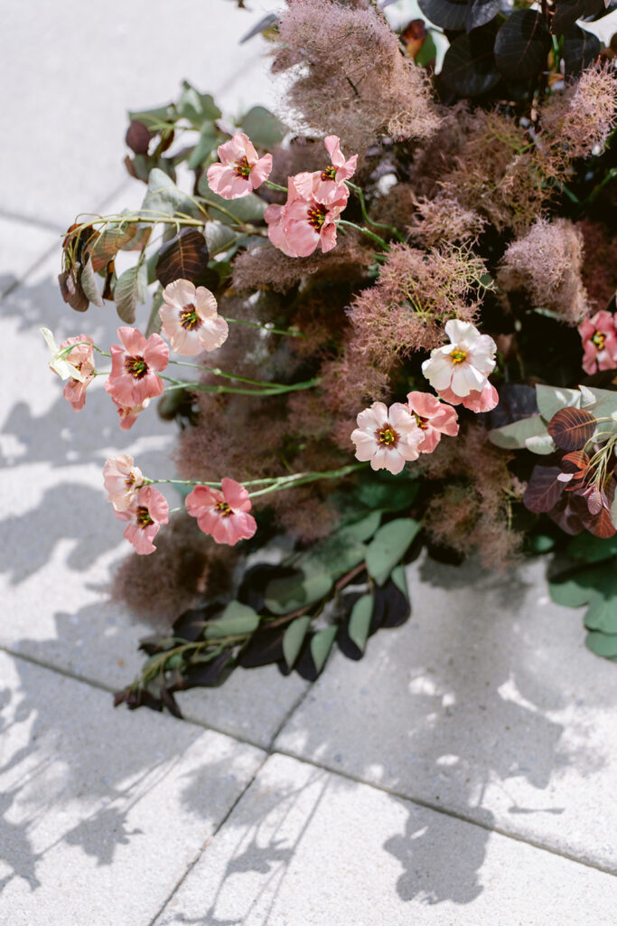 Close-up of pink cosmos and smoke bush blooms in a textured floral arrangement, casting soft shadows on stone paving, styled by Amelia at A. Carleton Weddings.