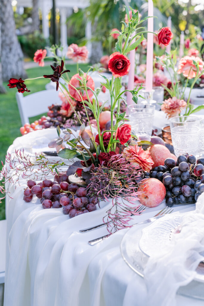 Wedding tablescape with red and pink floral arrangements paired with grapes and pomegranates, styled atop a flowing white linen for a rich, fruit-accented centerpiece.