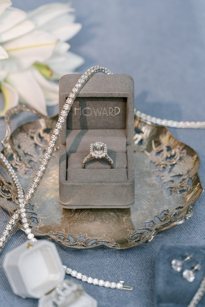 Diamond engagement ring in a velvet box labeled "Howard," styled with pearl bracelets and earrings on an antique silver tray.