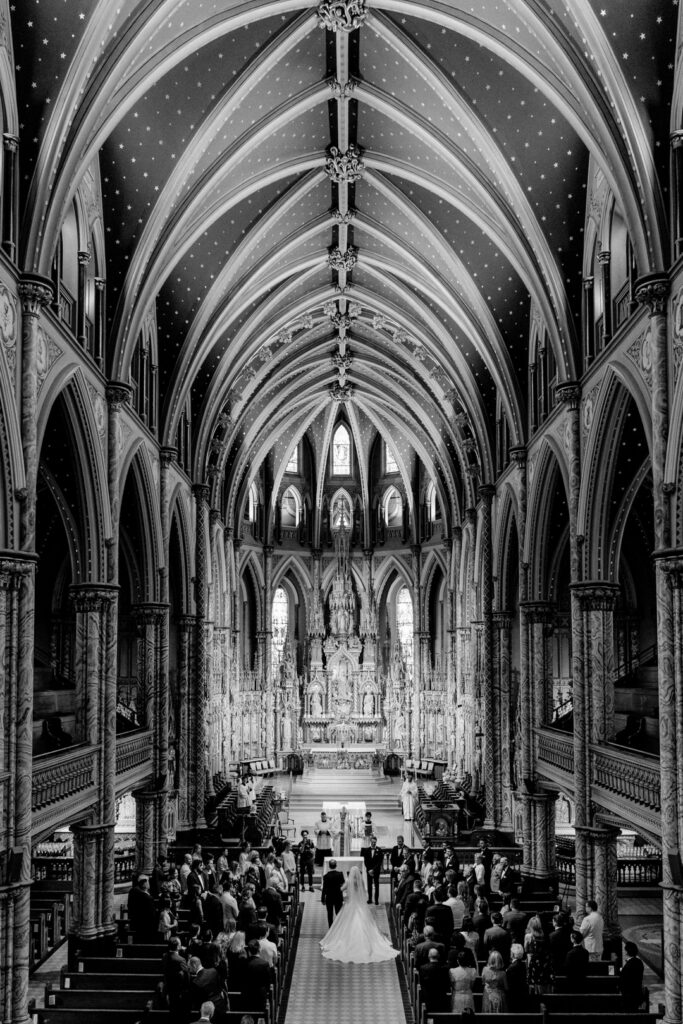 Black-and-white photo of a Gothic cathedral interior during a wedding ceremony, with soaring vaulted ceilings and guests seated in the pews below.