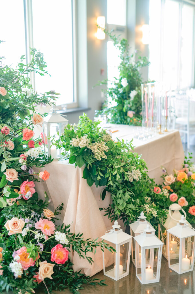 Lush greenery and pink-and-peach floral garland draping the head table, with white lanterns lining the bases, planned by Amanda from Sea and Silk Events
