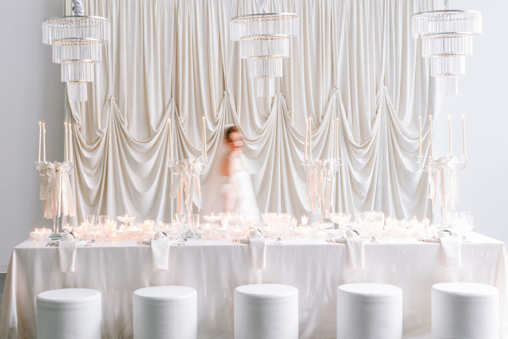 Motion-blurred bride walking behind an all-white candlelit reception table, set beneath tiered crystal chandeliers and draped ivory fabric.