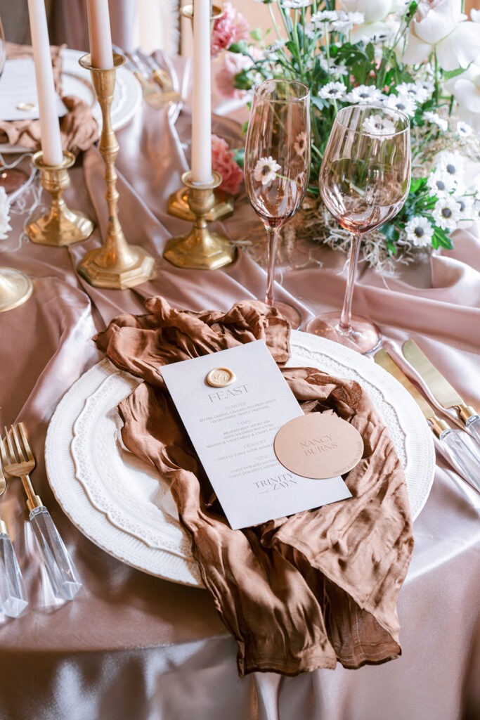 Elegant place setting with a bronze silk napkin, wax-sealed menu card, gold candlesticks, and smoked glassware, framed by white daisies.