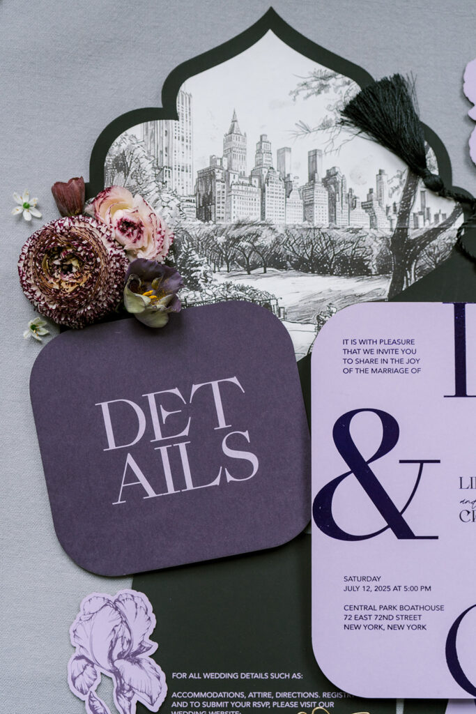 Layered wedding invitation suite in moody purple and charcoal tones, featuring a scalloped detail card, illustrated city skyline, tasseled tag, and dried floral accents.