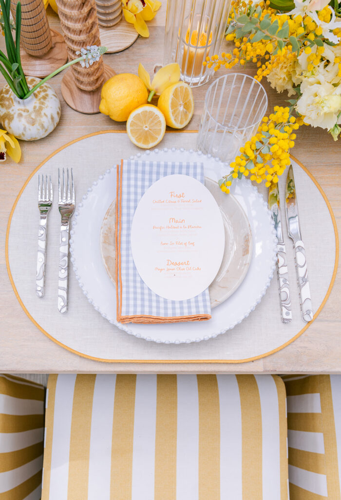 Sunny wedding place setting with a scalloped white plate, blue-and-white gingham napkin, printed menu card, textured silverware, and yellow floral and citrus accents.