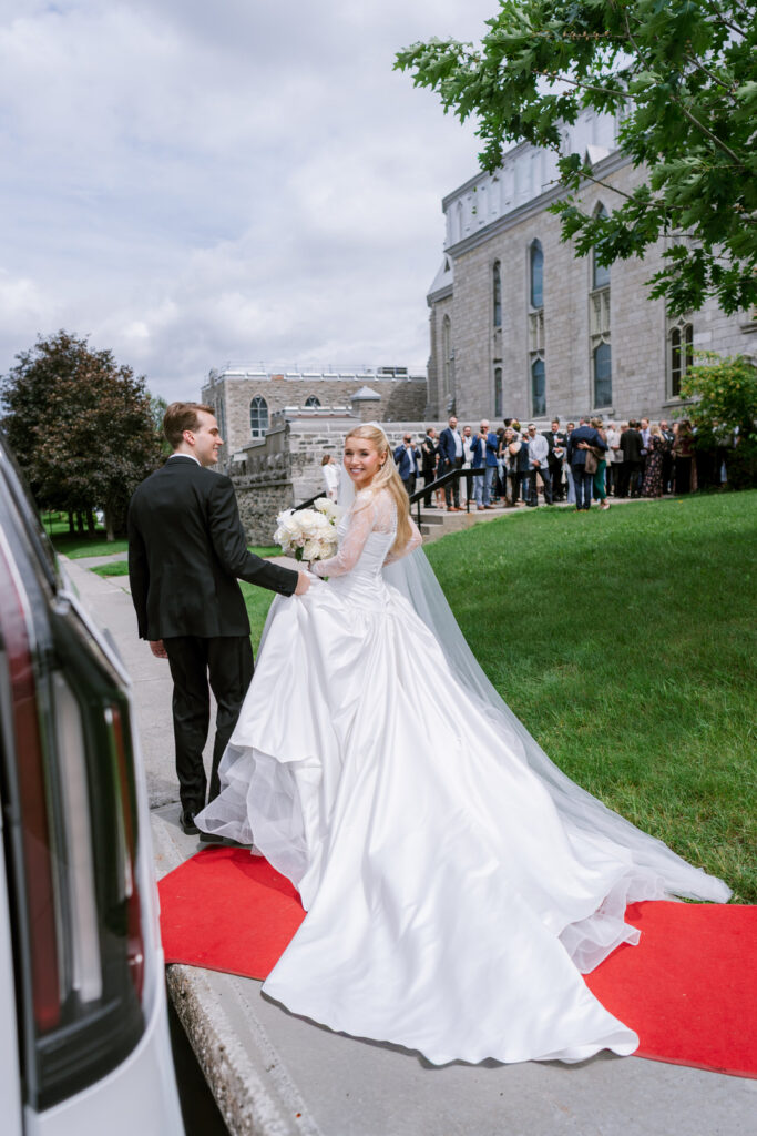 Bride and groom walking down a red carpet toward a stone building, guests gathered on the lawn in the background.