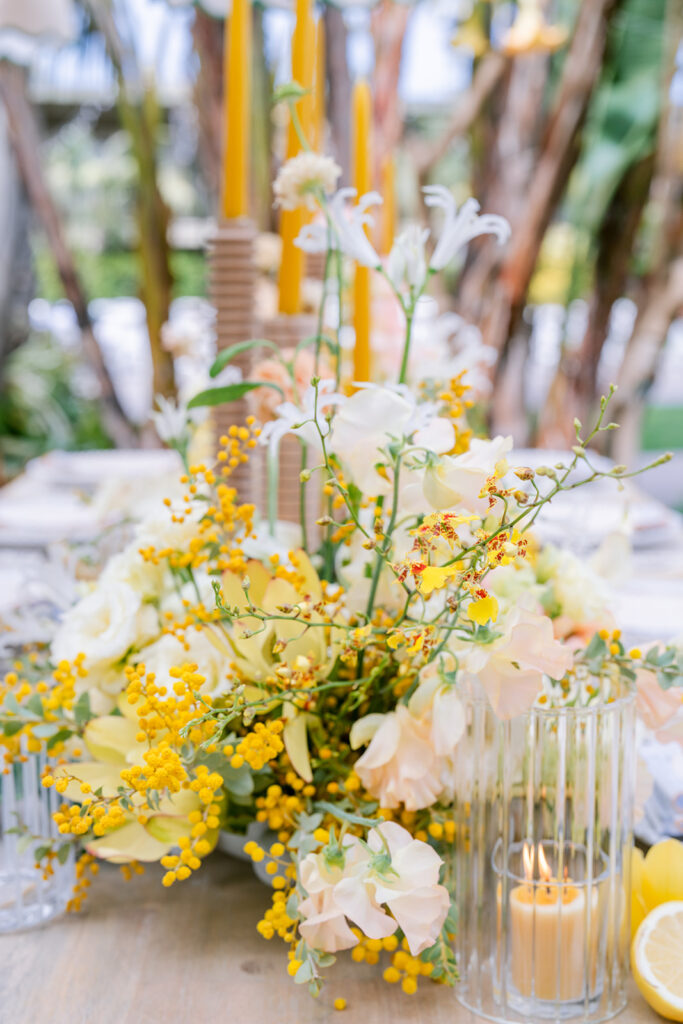 Close-up of a wedding table centerpiece with white and yellow blooms, mimosa sprigs, and tall mustard-hued taper candles among glassware.