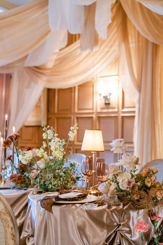 Warm, romantic table setting draped in champagne fabric, styled with a vintage lamp, taper candles, and lush floral arrangements.