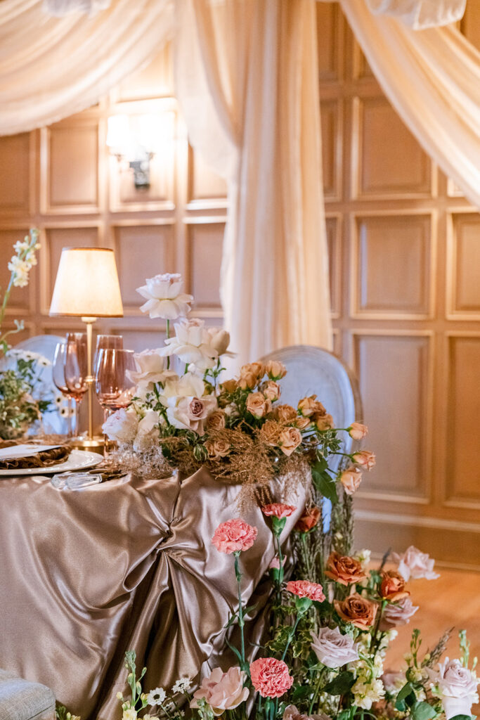 Close-up of a floral-draped chair back and table setting with warm pink blooms and a vintage lamp, styled by Michael at Wedecor.
