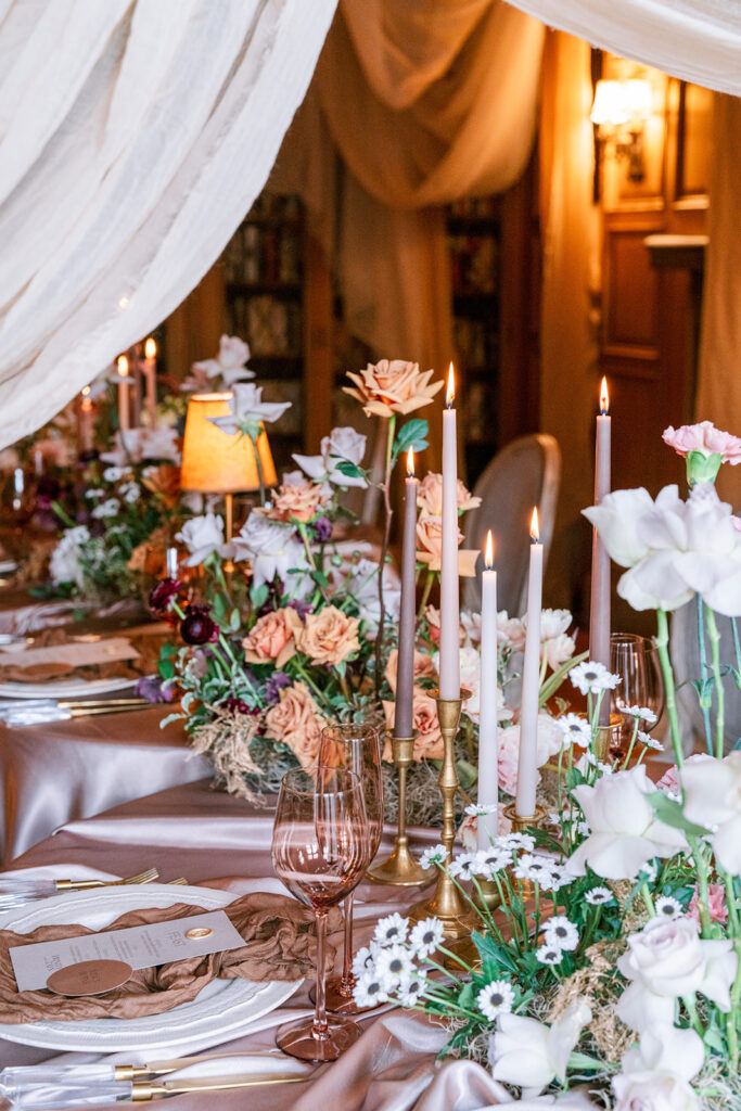 Long candlelit reception table dressed in warm mauve linens, with lush mixed floral arrangements, taper candles, and draped fabric overhead in a softly lit, romantic setting.