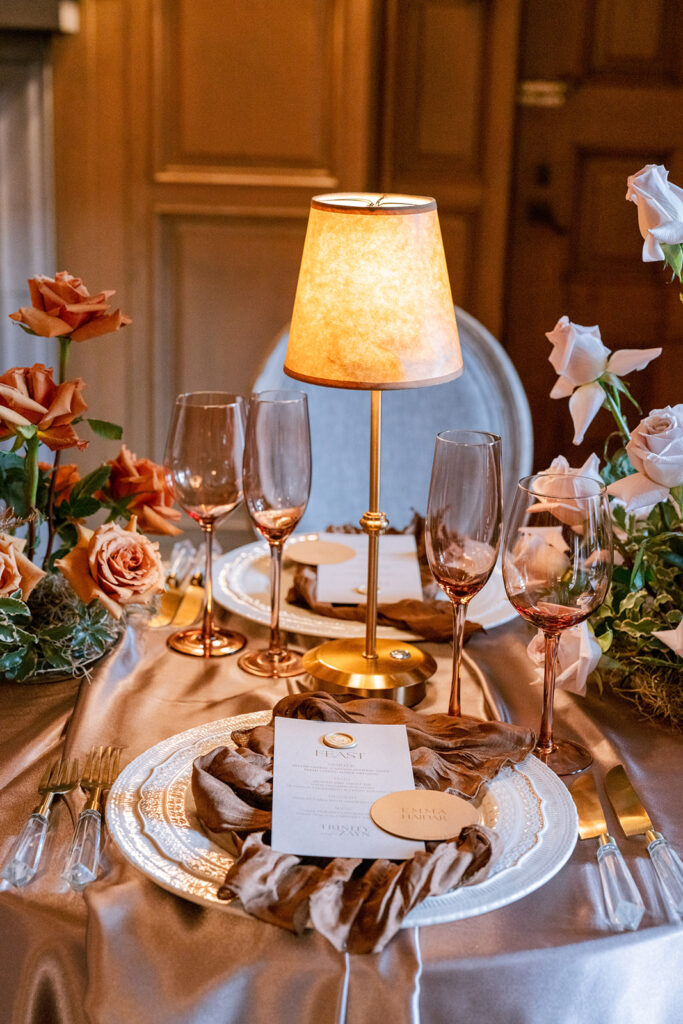 Warmly lit wedding place setting with a vintage-style table lamp, smoked glassware, gold flatware, and roses on a champagne satin linen.