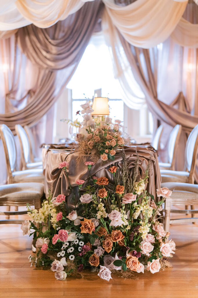Sweetheart table cascading with lush pink and mauve florals, framed by champagne draped fabric and a vintage-style lamp, styled by Michael at Wedecor.