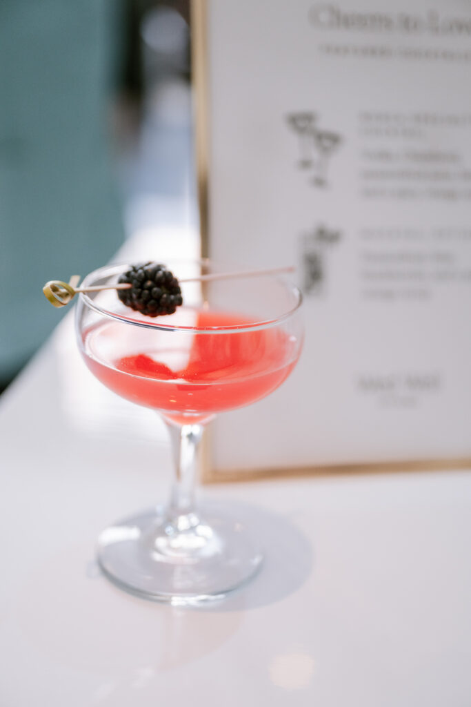 Pink signature wedding cocktail garnished with a blackberry, served beside a printed cocktail menu, styled by Amanda Simplice.