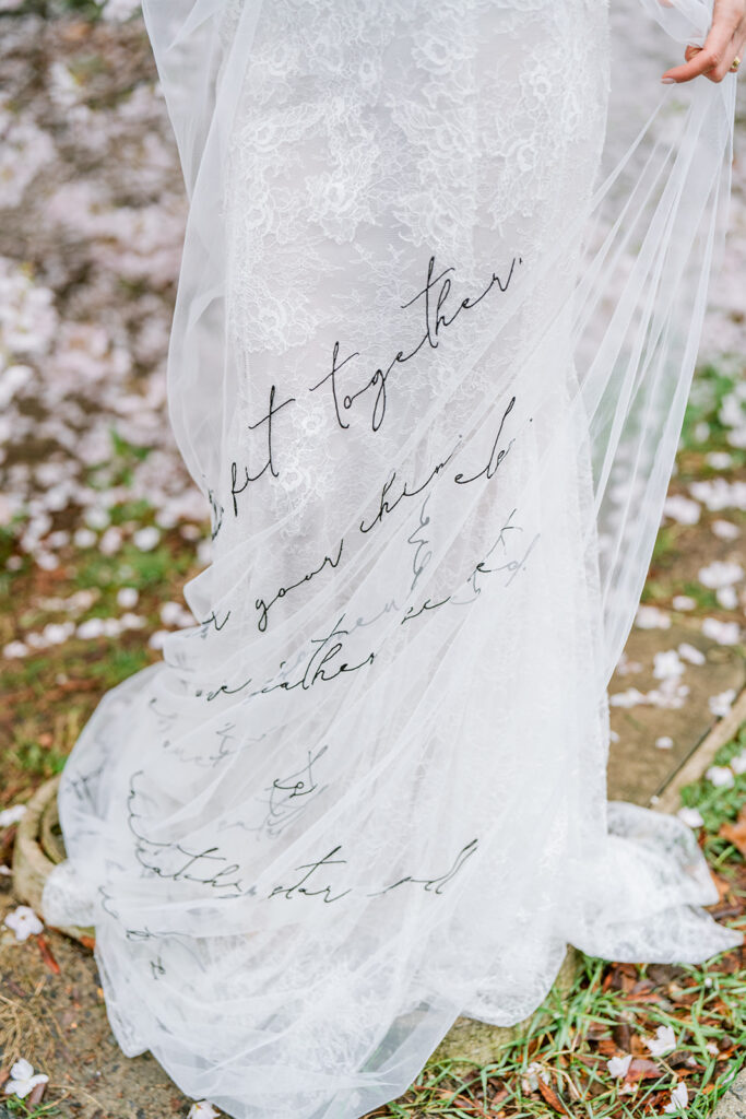 Close-up of a bridal veil embroidered with handwritten script, held up outdoors amid falling petals.