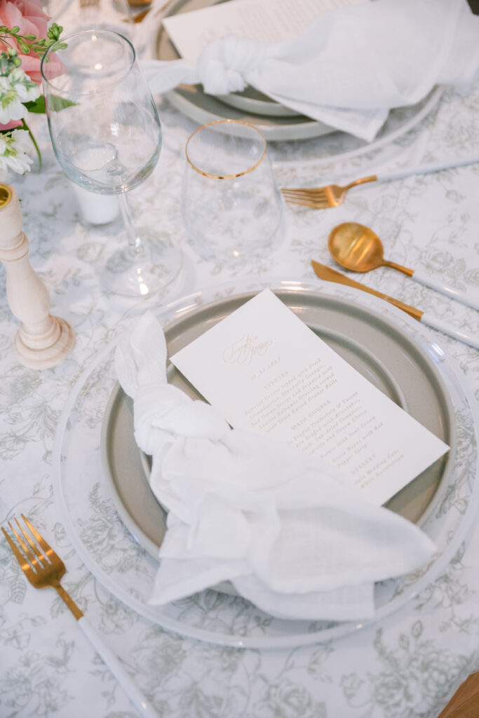Elegant place setting with a floral-patterned linen, gold flatware, and a monogrammed menu card, styled by Amanda Simplice.