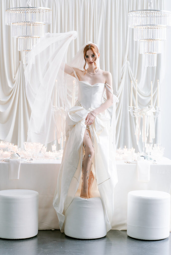 Bride in a strapless gown with a flowing veil, posed dramatically atop a white pedestal against draped fabric and hanging crystal fixtures, styled by Elise at Toast Events.