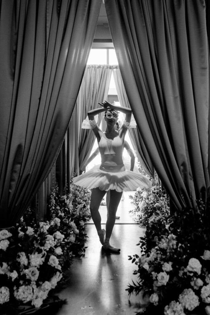 Black-and-white photo of a ballerina mid-pose in a draped doorway lined with white floral arrangements, styled by Amanda Simplice.
