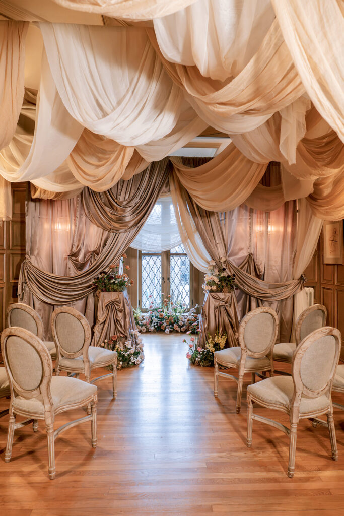 Wedding ceremony aisle draped in layered champagne and ivory fabric, lined with upholstered chairs leading to a floral-framed window.