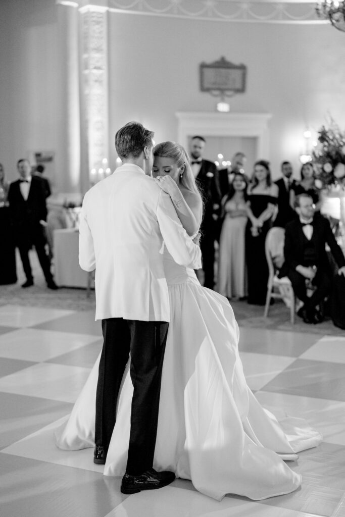 Black-and-white photo of a bride and groom's first dance, watched by guests in a grand ballroom with a checkered floor, styled by Erica Irwin Weddings & Events.