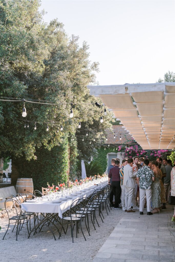 Guests gather beneath string lights and a pergola beside a long outdoor dinner table, styled by Amelia at A. Carleton Weddings.