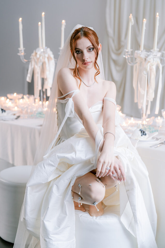Bride in a strapless gown crouching on a stool, styled against a soft backdrop with tall candelabras and draped fabric.