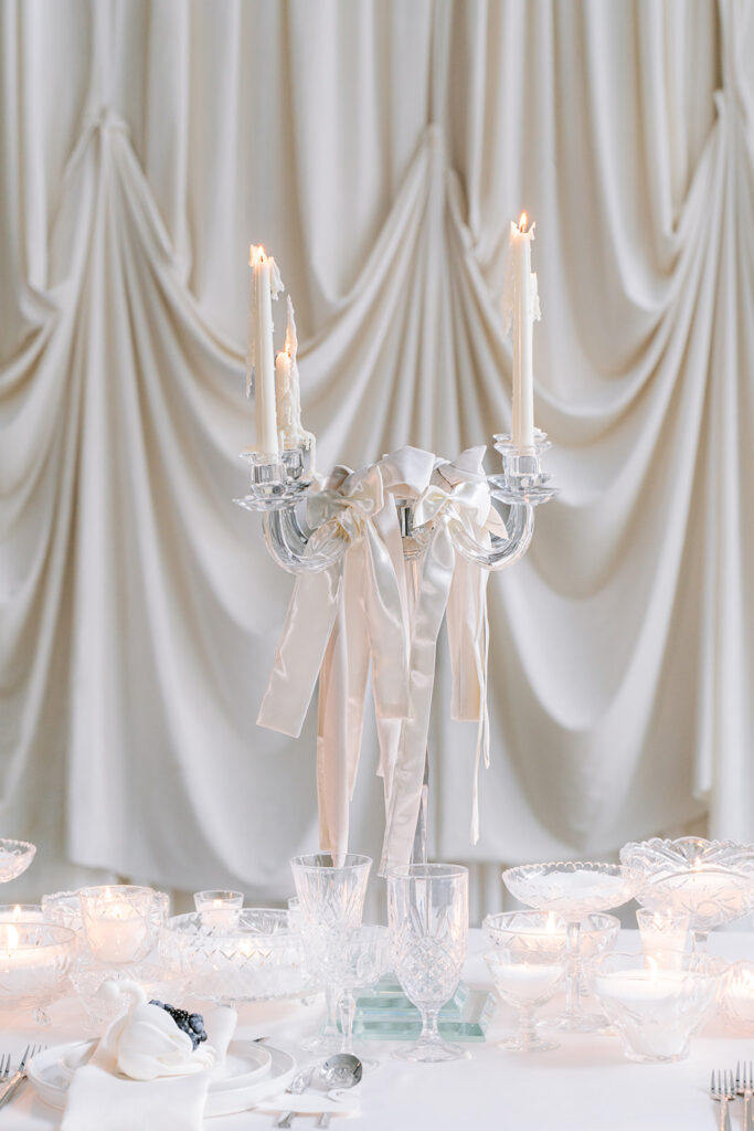 Candlelit tablescape with glass candelabras, ribbon accents, and delicate glassware set against draped ivory curtains, styled by Elise at Toast Events.