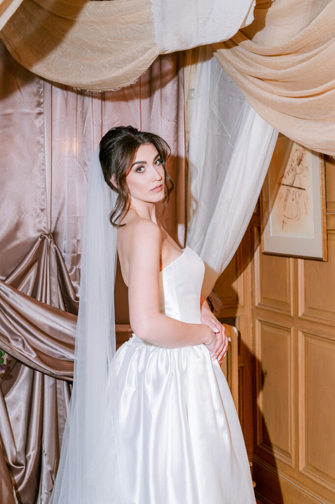 Bride in a strapless ballgown standing beside draped champagne fabric in a warmly lit interior.