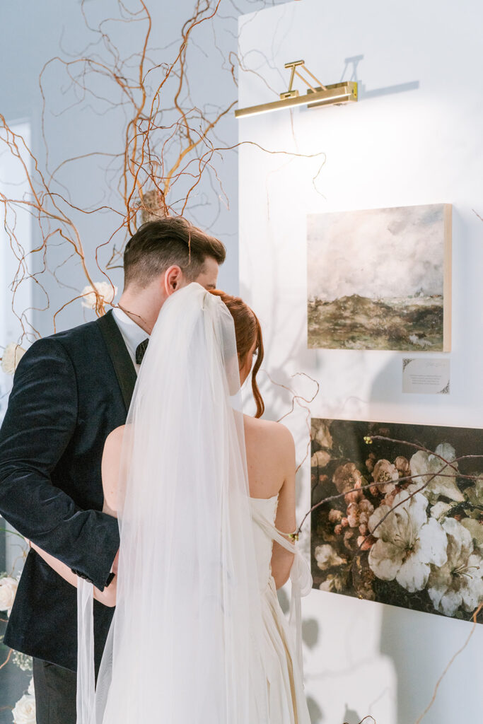 Bride and groom seen from behind, standing close together in front of framed artwork with a curly willow display above, styled by Elise at Toast Events.