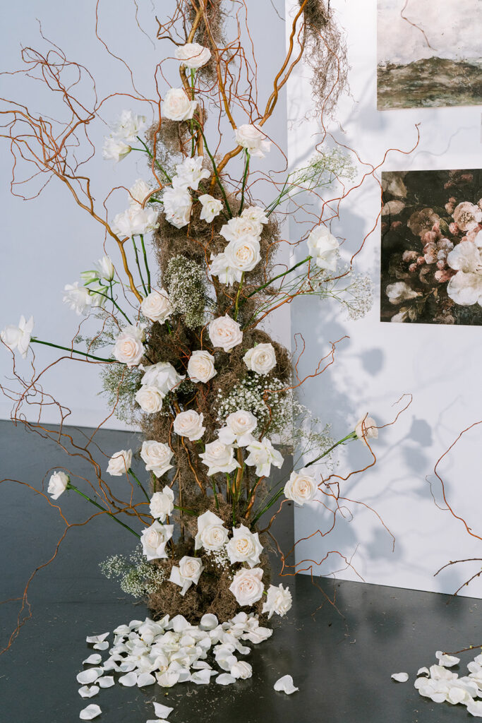 Sculptural wedding installation of white roses and baby's breath woven through curly willow branches, with scattered petals at the base and reference imagery visible behind it.