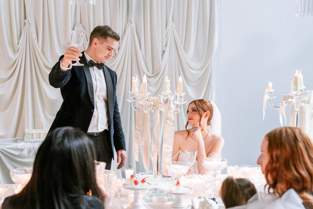 Groom raising a champagne glass in a toast beside a smiling bride at a candlelit reception table draped in white.