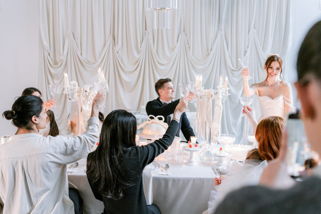 Bride and guests raising their glasses in a toast around a candlelit table, set against a backdrop of flowing draped white fabric.