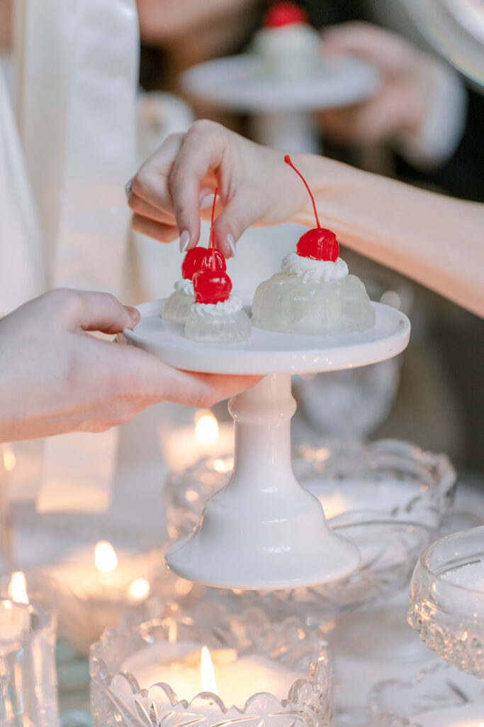 Close-up of hands placing cherry-topped mini desserts on a white pedestal cake stand, surrounded by glowing votive candles, styled by Elise at Toast Events.