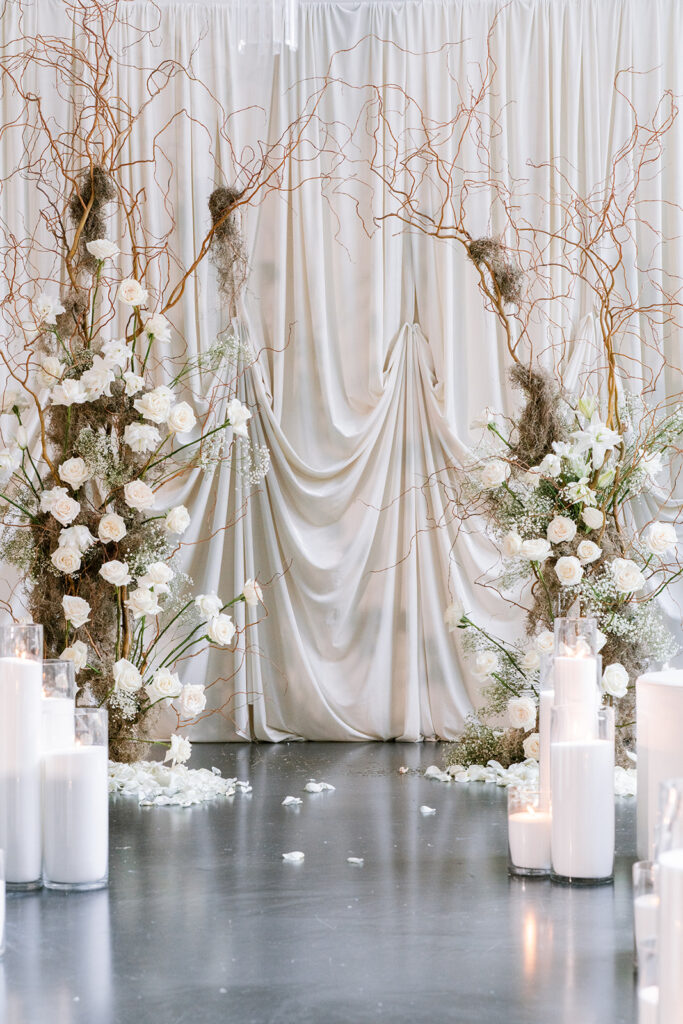 Ceremony backdrop of draped ivory fabric with white rose and branch arrangements and pillar candles, from a Rococo-inspired editorial at the Ottawa Art Gallery by Elise of Toast Events, Ottawa wedding planner.