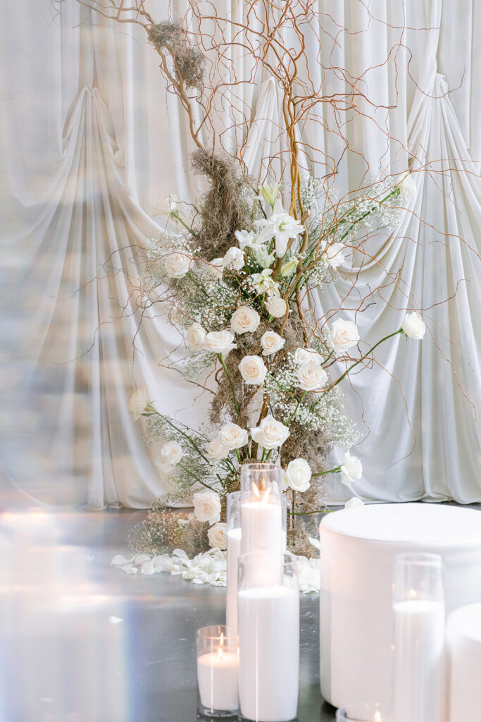 Ethereal wedding installation of white roses and curly willow branches rising above pillar candles, set against soft draped ivory fabric.