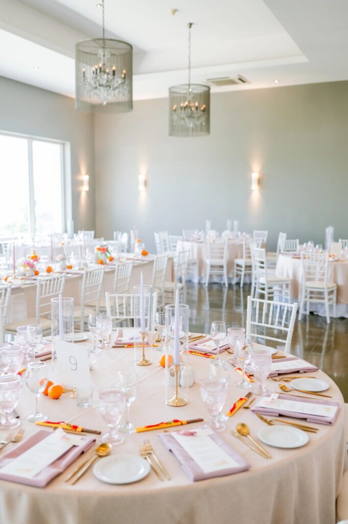 Wedding reception room set with round tables, white chiavari chairs, and pendant chandeliers under soft natural lights, planned by Amanda from Sea and Silk Events
