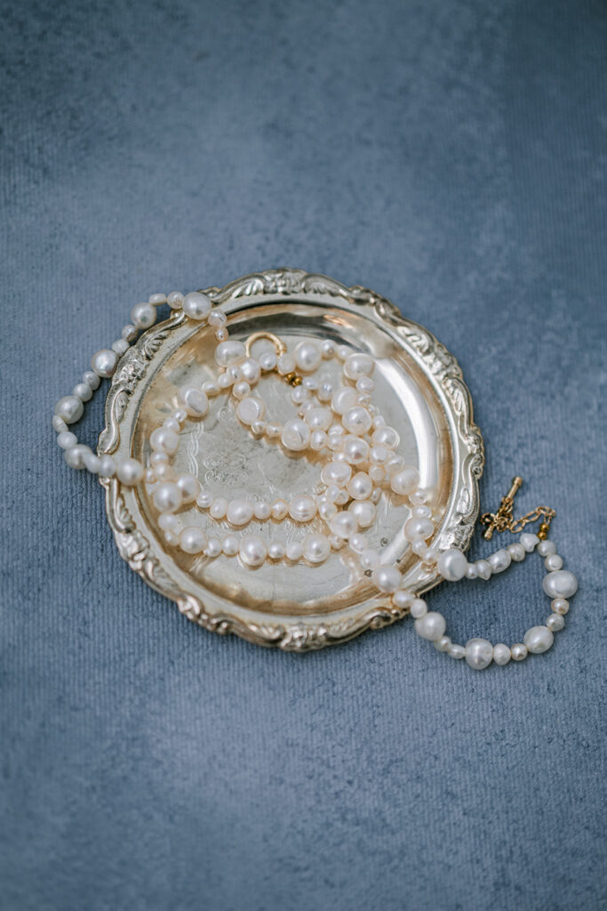 Vintage pearl necklaces and gold jewelry displayed on an ornate silver tray against a dusty blue background.