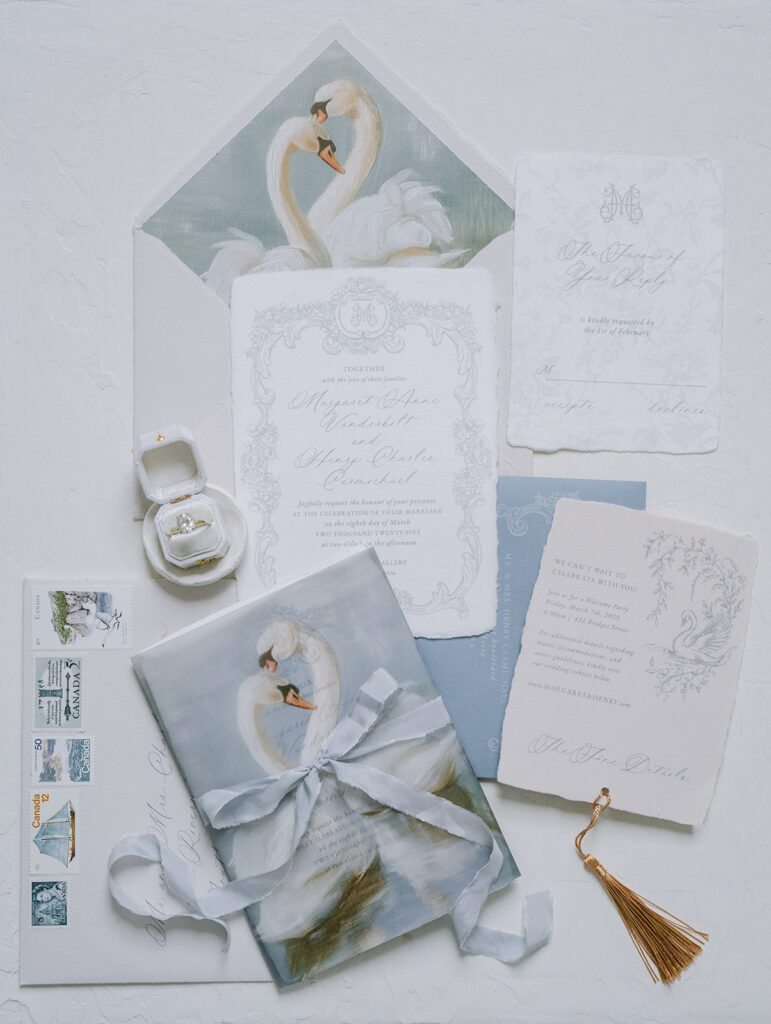 Romantic wedding invitation suite in soft blue and white, featuring an ornate scalloped invitation, swan-illustrated envelope liner and wrap, gold tassel, and vintage postage stamps.
