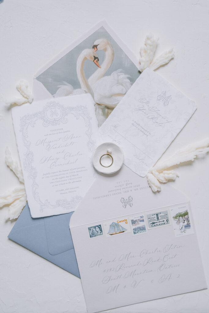 Wedding invitation suite in dusty blue and white, with a swan-illustrated envelope liner, ornate scalloped card, ring box, and vintage postage stamps.