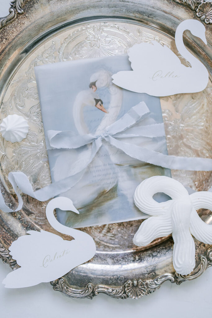 Wedding invitation wrapped in a silk ribbon bow atop an antique silver tray, styled with paper swan cutouts and a decorative shell.