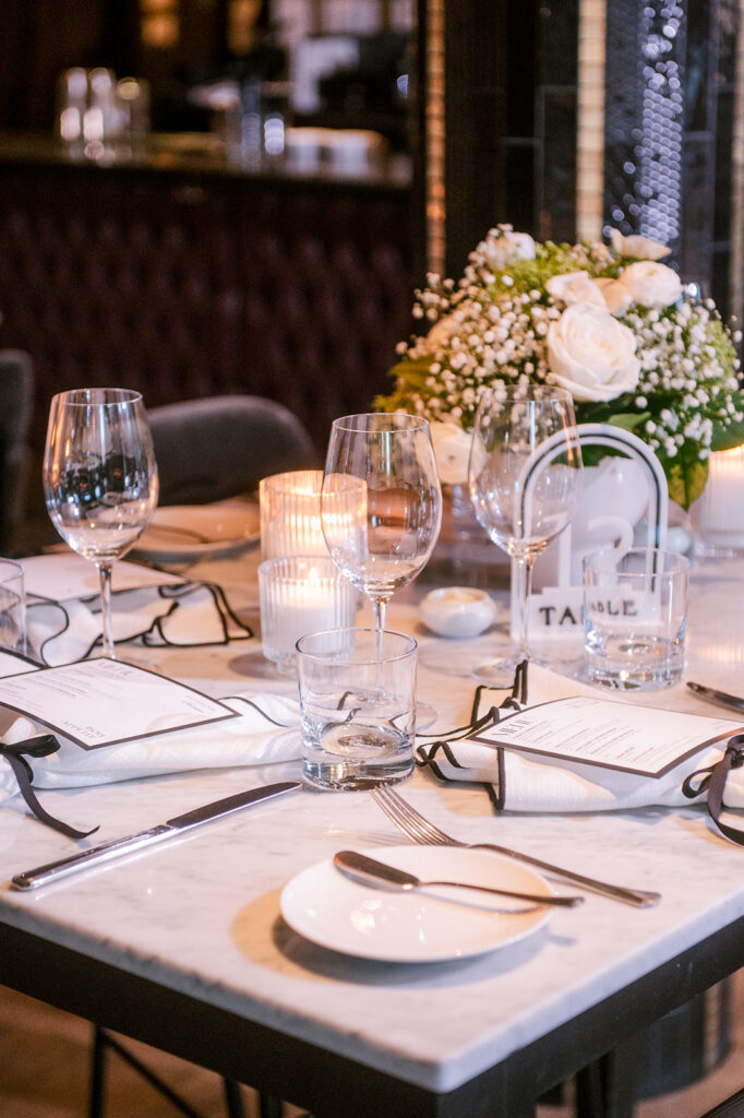 Close-up of a marble table setting with a hanging glass table number, white floral centerpiece, and ribbon-tied menu card, styled by Cindy and Rita at All In Event Planning & Design.