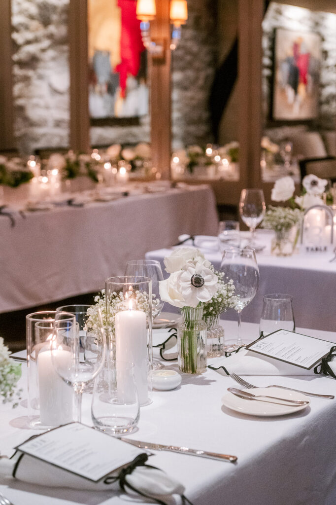 Long candlelit reception table with white anemone centerpieces, ribbon-tied menus, and pillar candles in a stone-walled room, styled by Cindy and Rita at All In Event Planning & Design.