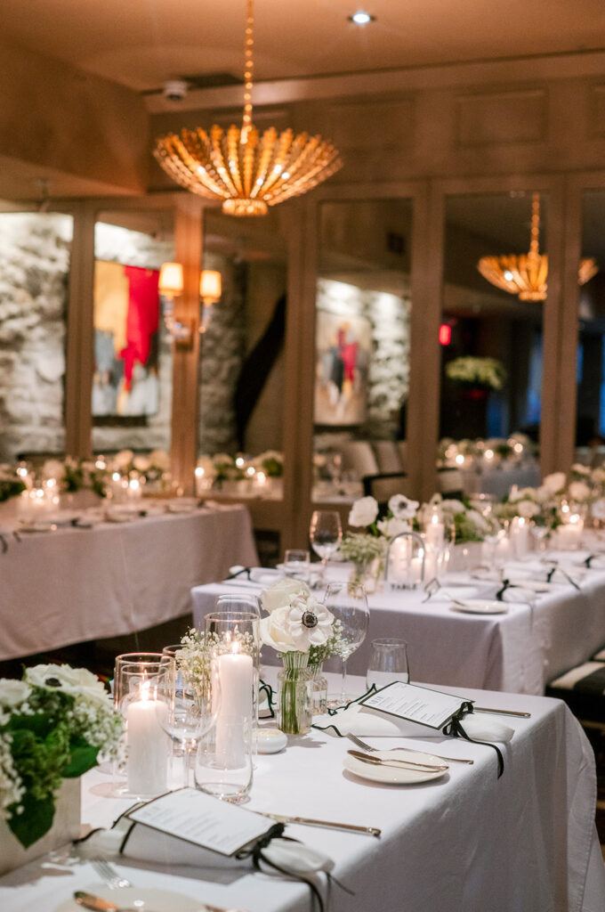 Long candlelit reception tables set with white florals and menu cards beneath a gold pendant chandelier in a stone-walled dining room, styled by Cindy and Rita at All In Event Planning & Design.